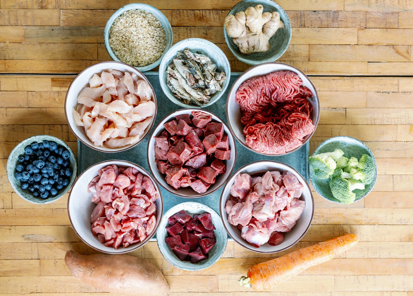 Bowls of fresh chicken, beef, turkey, liver, sardines, oats, ginger, blueberries and broccoli arranged on a wooden board, with sweet potato and carrot alongside
