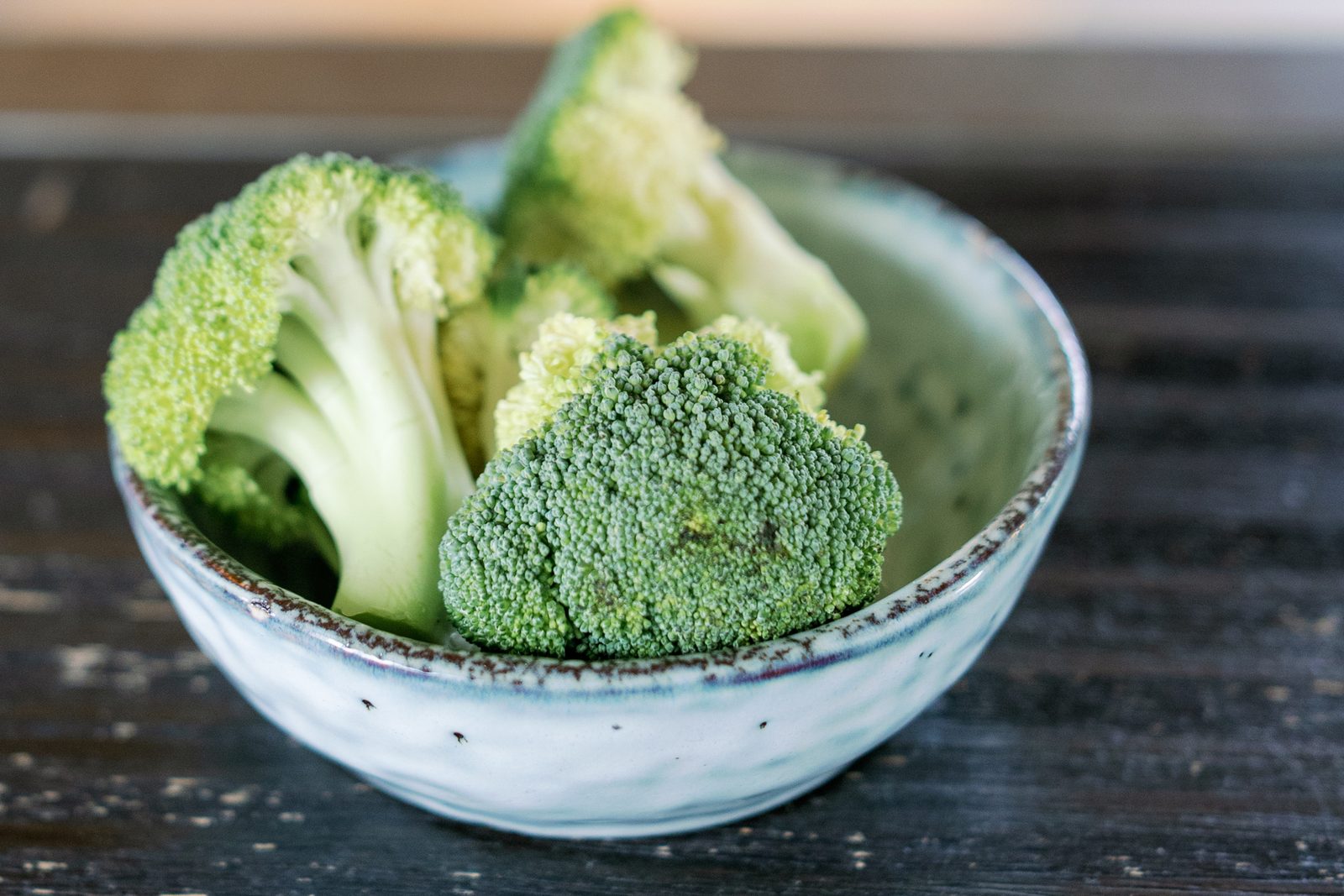 A small ceramic bowl of fresh broccoli florets on a dark wooden table