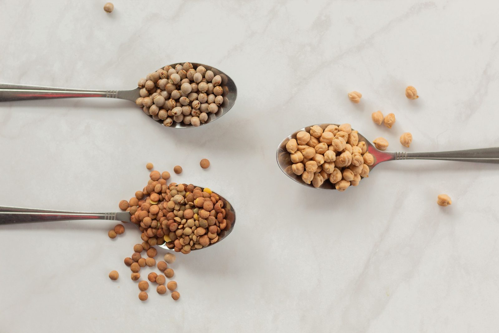 Three spoons of dried chickpeas, peas and lentils laid out on a white marble surface