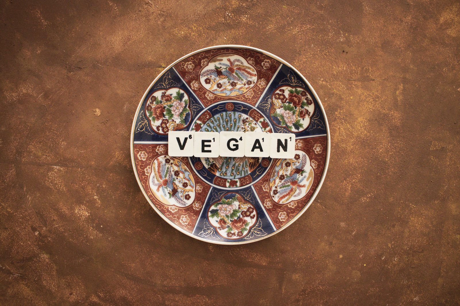 A decorated porcelain plate on a warm brown surface, with letter tiles on it spelling the word VEGAN